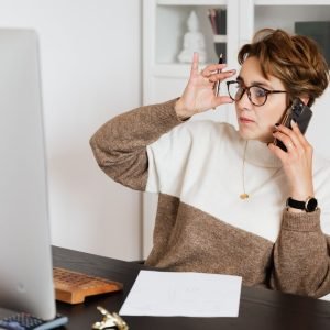 Serious good looking woman in glasses talking on smartphone and looking at computer screen with pen in hand and sheet of paper on desk